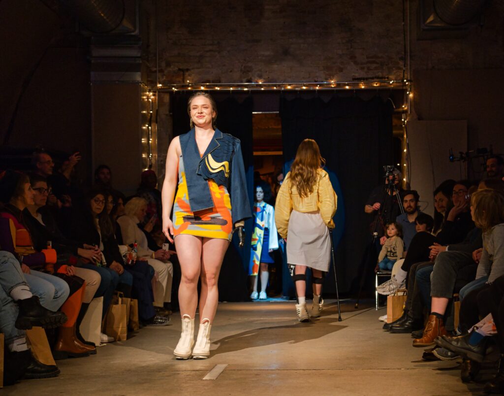 Models auf dem Laufsteg, vorn ein Model in buntem kurzen Kleid, blauer Jacke und weißen Stiefeletten