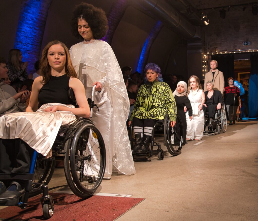 viele verschiedene Models in einer Reihe auf dem Laufsteg, u.a. Rolli-Models, Kleinwüchsige, ein blindes Model ...