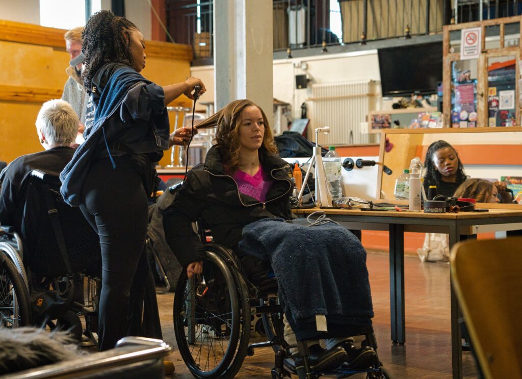 Backstage einer Modenschau, 2 Models in Rollies am Schminktisch, eine junge Frau wird gerade frisiert