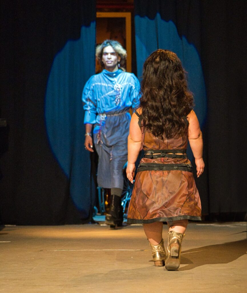 Buntfoto von einer Modenschau. im Vordergrund ein kleinwüchsiges Model im braunen Kleid, von hinten zu sehen, im Hintergrund eine groß gewachsene Person im blauen Kleid, durch ein Spotlight angeleuchtet.