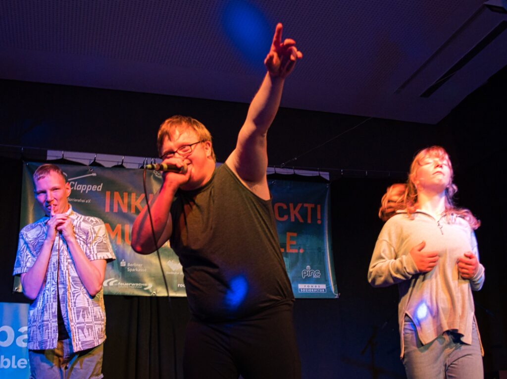 Ein Junger Mann singt auf der Bühne in ein Mikro und streckt den Arm zum Publikum aus.  daneben stehen zwei Jugendliche