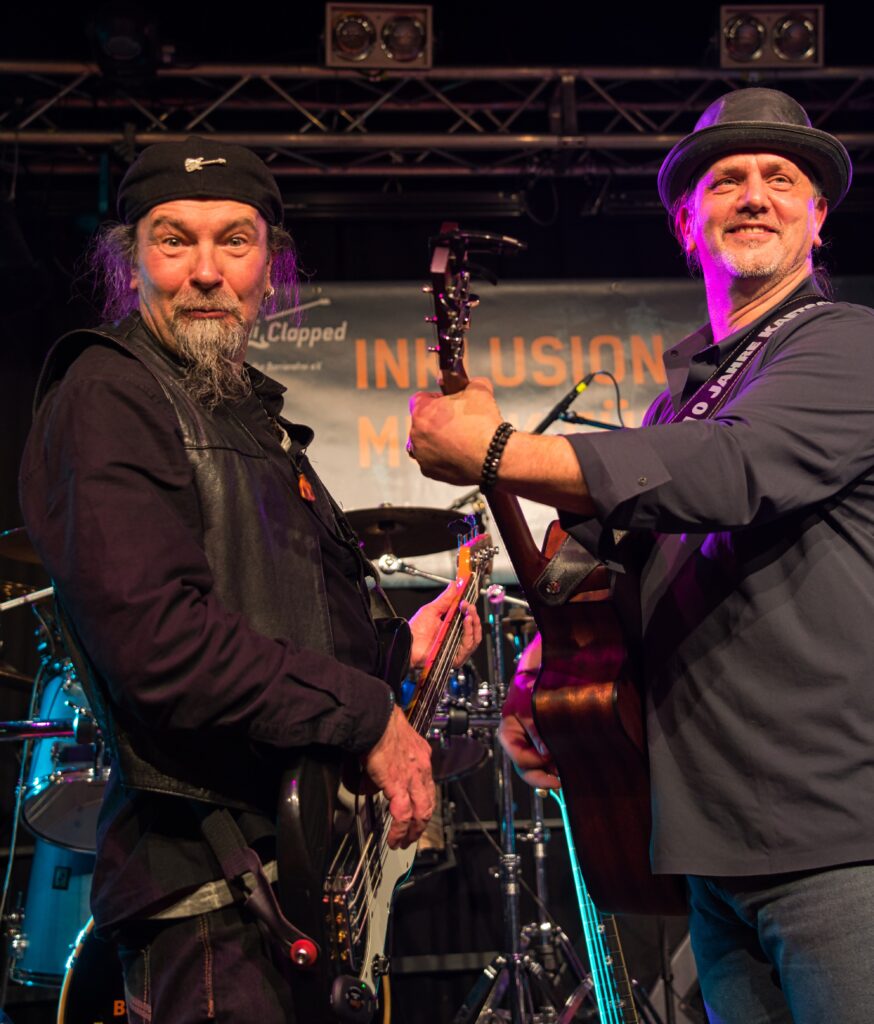 2 Gitarristen stehen sich gegenüber und spielen. Sie sehen zur Seite, direkt in die Kamera bzw ins Publikum