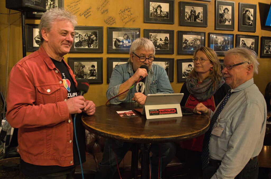 Peter, Jana und Achim von Handiclapped stehen mit Klaus von Rockradio an einem runden Kneipentisch Klaus spricht in ein Mikro, vor ihm auf dem Tisch ein Laptop