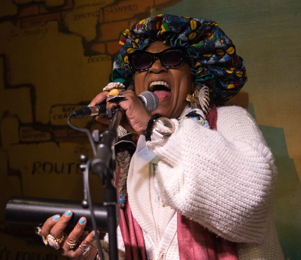 eine Sängerin am Mikrophon singt sehr kraftvoll. In einer Hand hält sie eine Rassel, sie trägt an allen Fingern Ringe, eine bunte Mütze und ein dunkle Brille