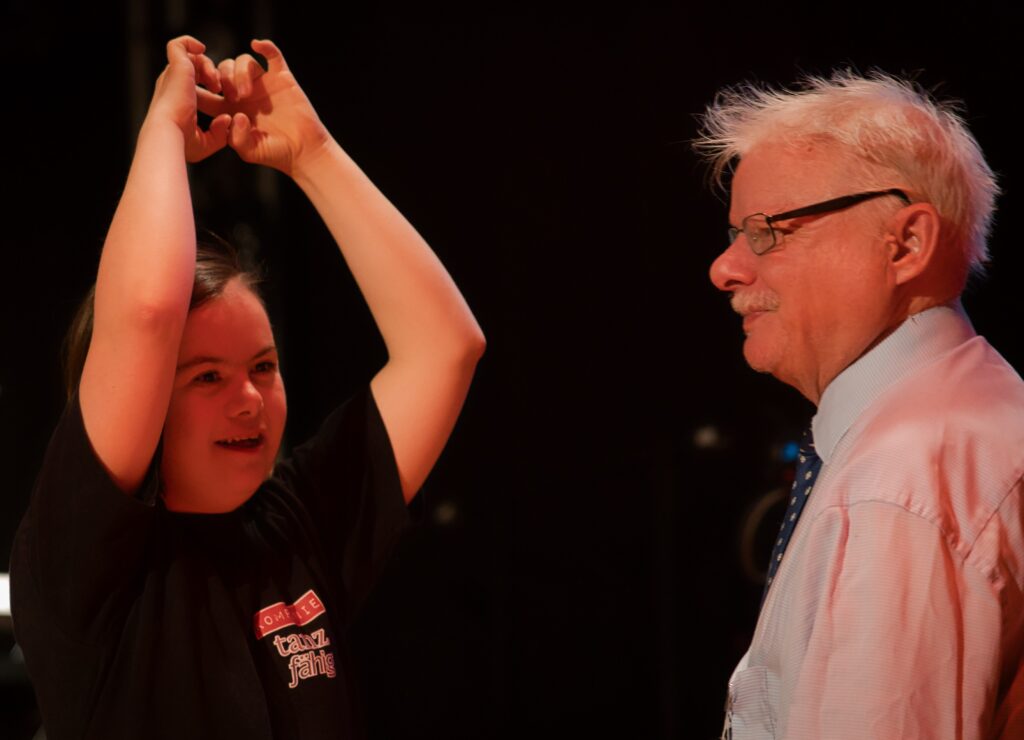 Buntfoto von zwei Menschen, rechts Achim und links eine junge Frau,die die Arme gen Decke streckt.