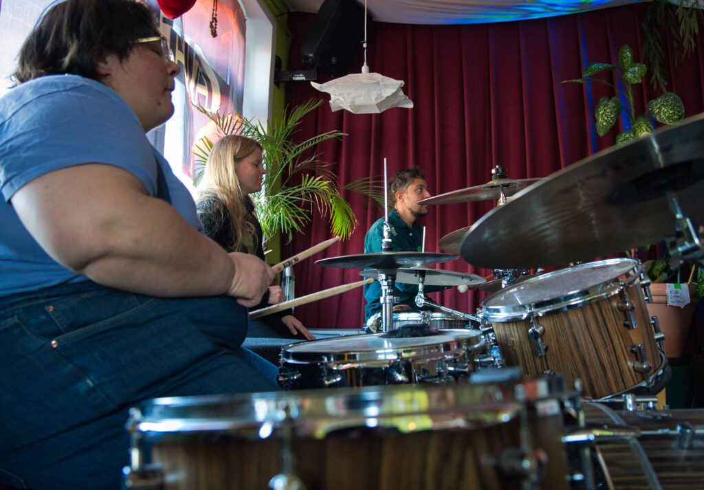 2 Frauen und ein Mann sitzen in einem Studio jeweils an einem Schlagzeug und spielen zusammen
