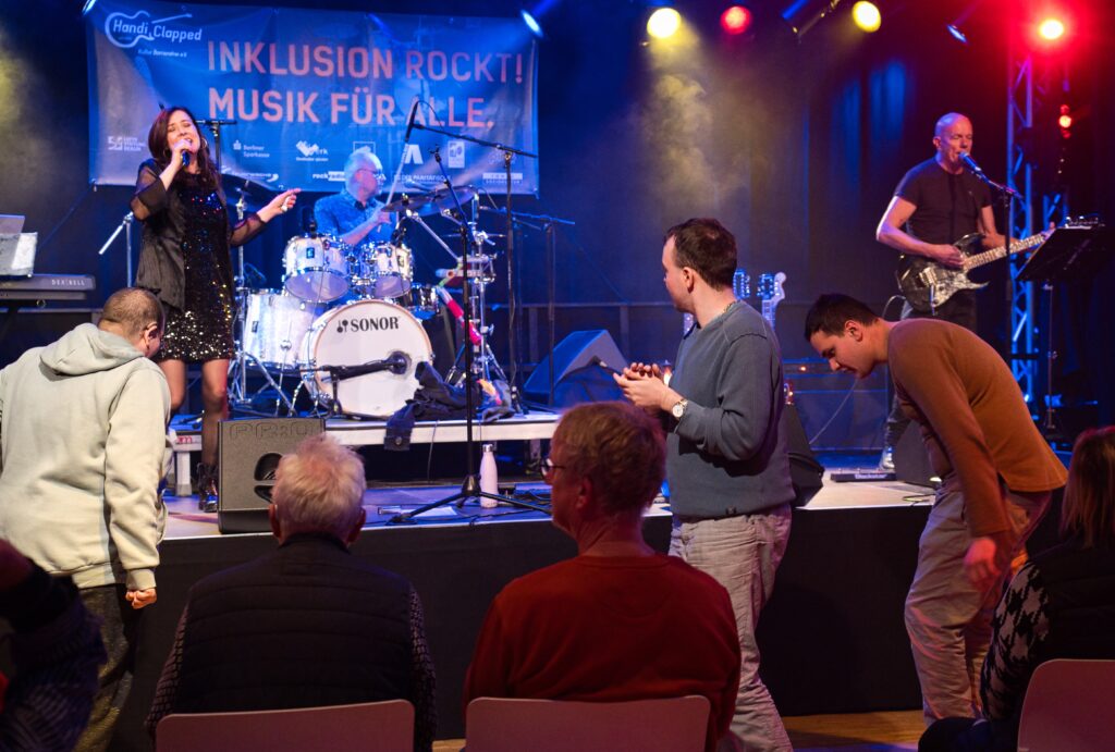 eine Band mit Sängerin, Schlagzeuger und Gitarrist auf der Bühne. Vor der Bühne tanzen 3 junge Männer