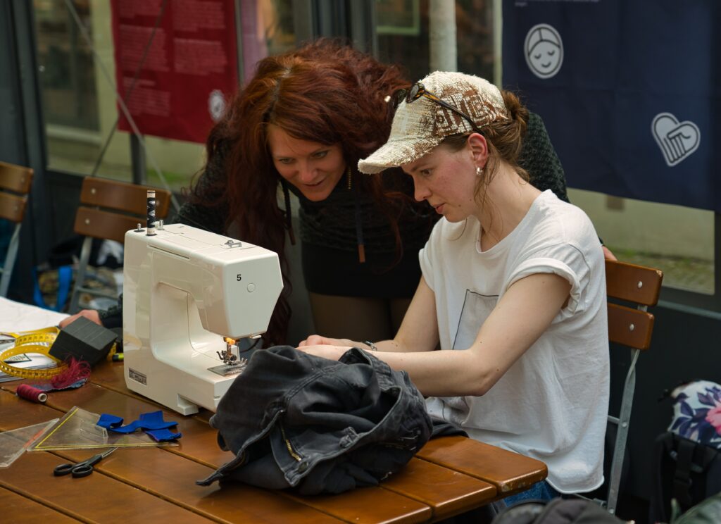 eine Frau an einer Nähmaschine, eine andere Frau sieht ihr interessiert auf die Hände