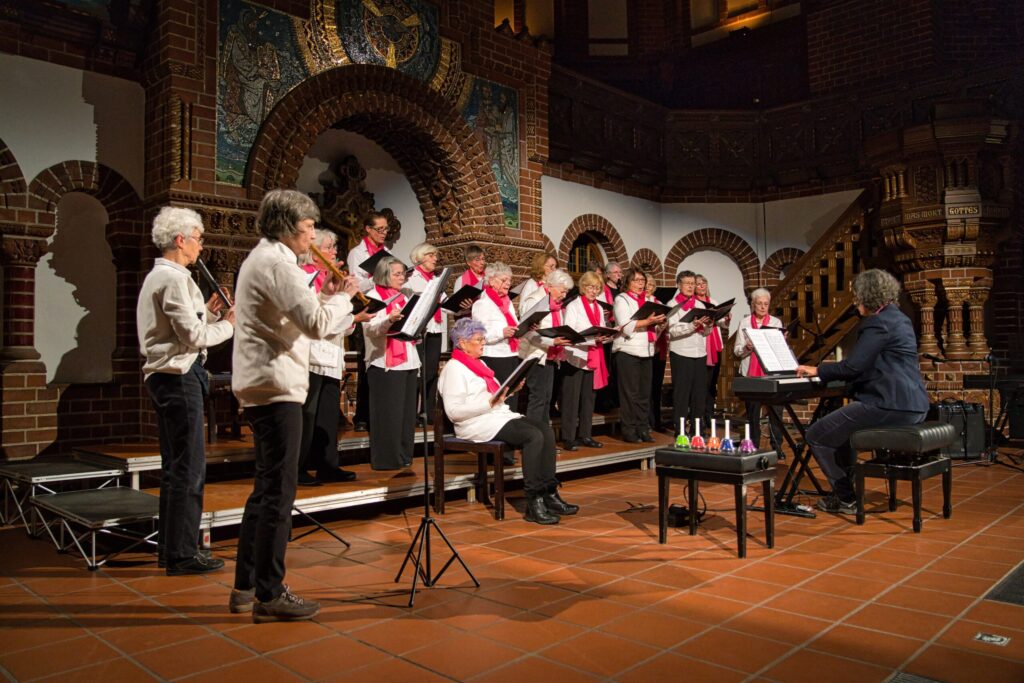 Ein Chor von ca- 20 Frauen die in einer Kirche singen, eine Spielt Keyboard und zwei Flöte. Alle tragen weiße Hemden und rosa Tücher. 
