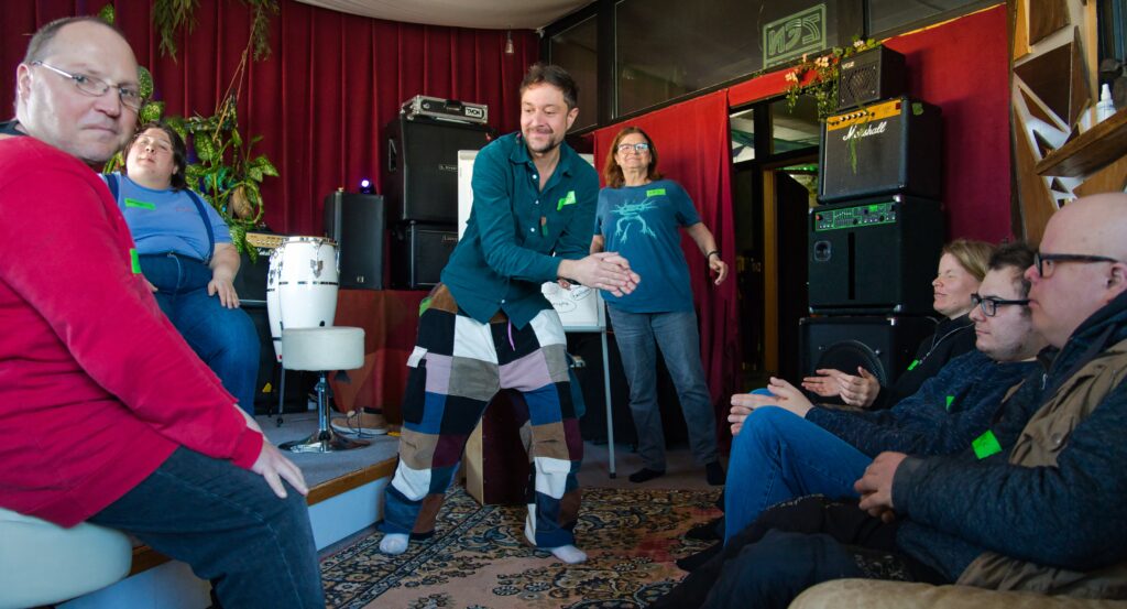 die Workshopteilnehmer sitzen und stehen im Kreis. Der Workshopleiter in der Mitte. Sie sind in einer Aufwärmübung durch Klatschen und BodyPercussion