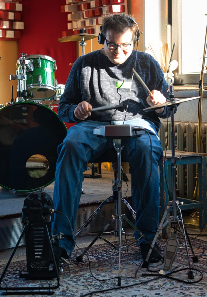 ein Workshopteilnehmer in einem Studio mit Kopfhörern. Er trommelt mit Stöcken auf E-Drums. Er lächelt dabei. 