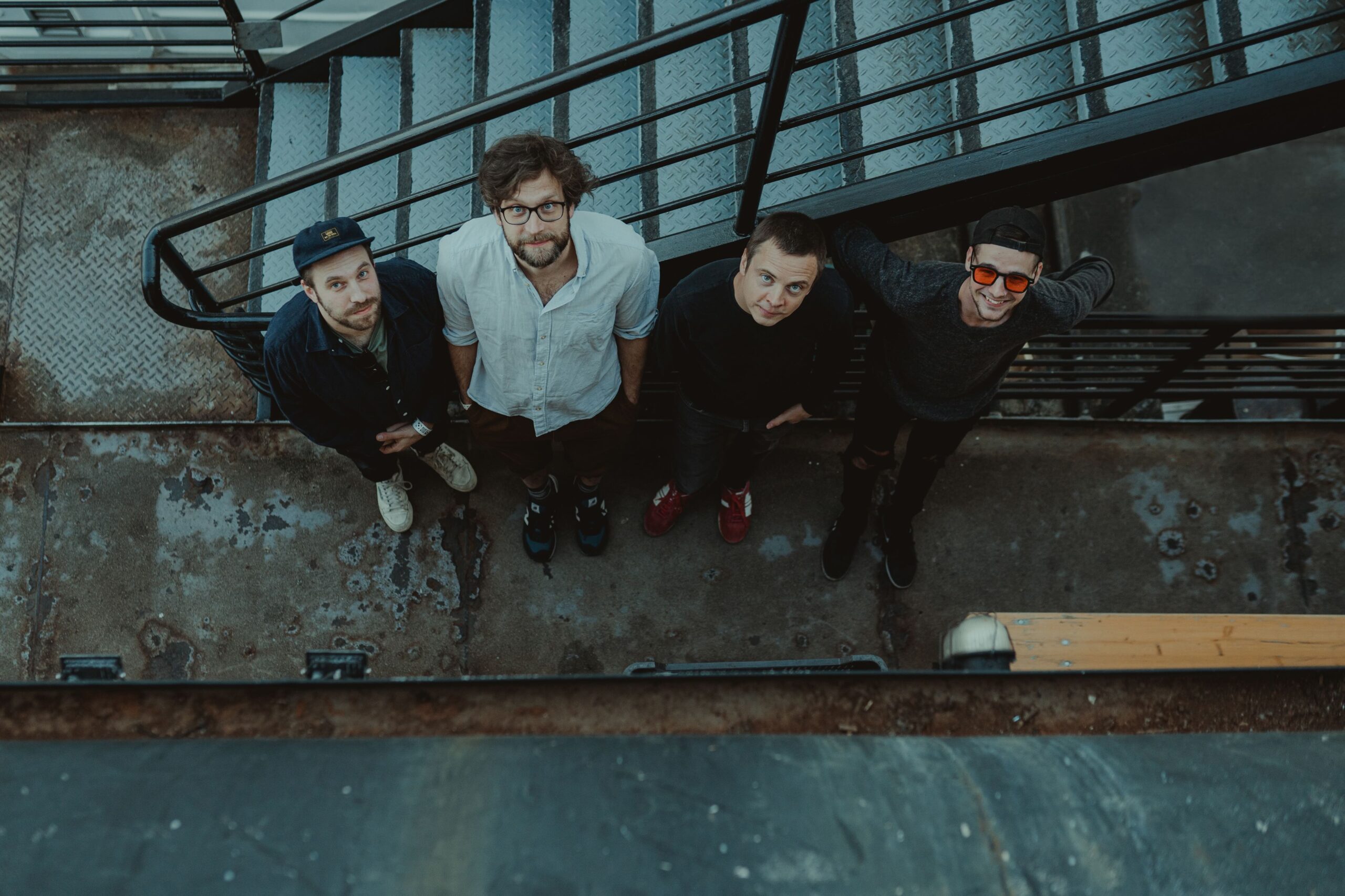 Buntfoto des Quartett. Sie stehen vor einerm Treppengeländer im Fruen, von oben Fotografiert und sie gucken nach oben in die Kamera