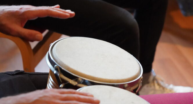 Es sind die Hände von zwei Personen beim Musizieren zu sehen. Die eine Person schlägt auf Bongos (kleine Trommeln). Die andere hält ein kleines Percussion-Instrument aus dunklem Holz in den Händen.