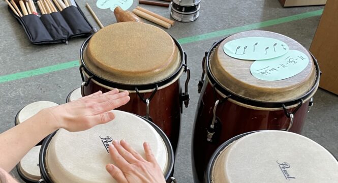 Eine Workshopsituation. Zwei Hände schlagen auf eine von vier Congas. Im Bild sind verschiedene Schlegel in einer Tasche und Moderationskarten mit Noten zu sehen.