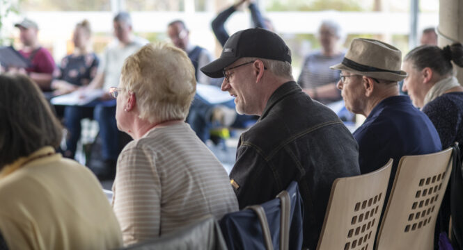 Viele Menschen unterschiedlichen Alter und Geschlechts sitzen in einem großen Stuhlkreis in einem hellen Raum. Die Gesichter sind fast alle unscharf. Ein Mann mit Kappe lächelt. Ein Mensch hat seine Arme nach oben gerissen und klatscht. Einige haben A4-Papier auf ihrem Schoß.