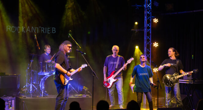 Buntfoto einer fünfköpfigen Band bei einem Auftritt auf einer Bühne. von links nach rechts: Schlagzeuer, E-Gitarriste, E-Bassist, Sängerin, E-Bassist, dazu dunkle Bühnenbeleuchtung und ein Schriftzug im Hintergrund: "Rockantrieb"