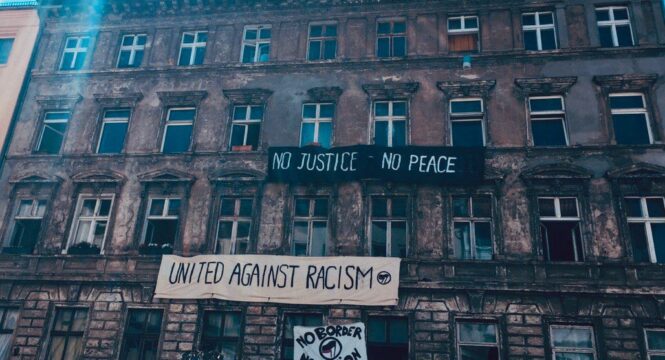 Ein verwittertes mehrstöckiges Wohnhaus mit mehreren Bannern an der Fassade, darunter Aufschriften wie "o Justice -No Peace", " United Against Racism" und "No Border - No Nation". Der Himmel über dem Haus ist blau und sonnig.