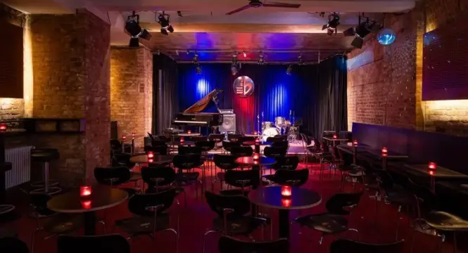 Buntfoto von einem Jazzclub im Leller, gedinegene Atmosphärre mit ca 15 Tsichen vor einer Bühne mit Flügel, SChlagzeug, Vrohang im Hintergrund und großem Blat Logo davor hängend.