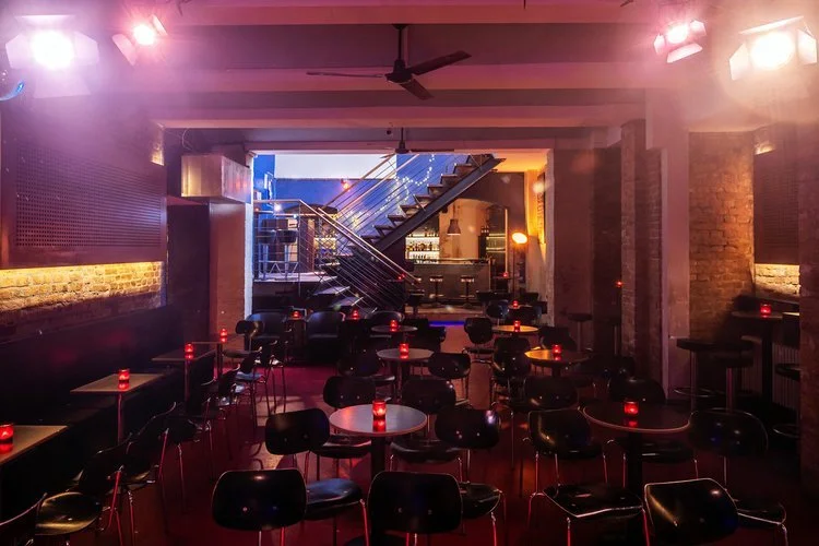 Foto von einem Raum im Keller mit ca 10 Tischen an denen je 2 Personen Platz haben, im Hintergrund eine Treppe nach oben und eine Bar