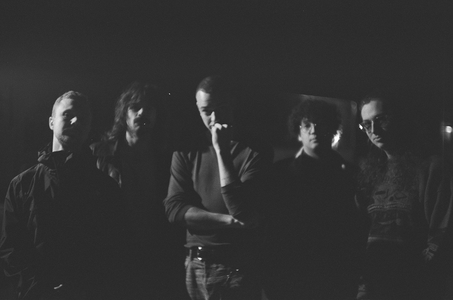 ein dunkles Foto in schwarz weiß zweigt fünf stehende, männlich gelesene Personen, die in die Kamera schauen. Sie haben unterschiedliche Frisuren. Eine trägt langes leicht gewelltes Haar und einen Schnauzbart. Die mittlere Person mit sehr kurzem Haar stützt mit zum Ellenbogen gewinkeltem Arm ihr Kinn.