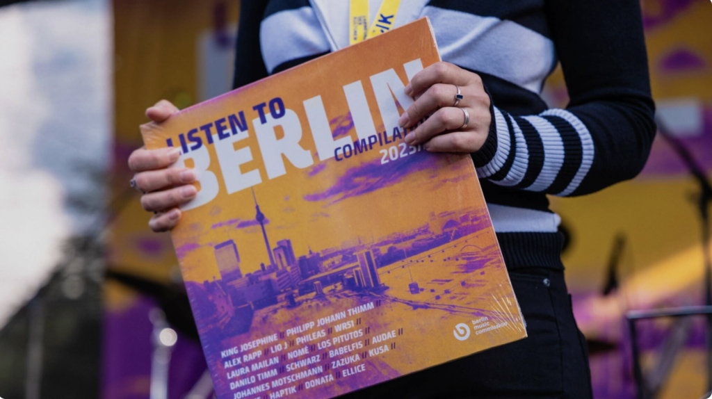 Buntfoto von einer Person die eine SChallplatte in den Händen vor ihren Körper hält. Darauf steht: Listen to Belrin und ein orange gefärbtes Bild von der Belrin Skyline ist auf dem Cover.