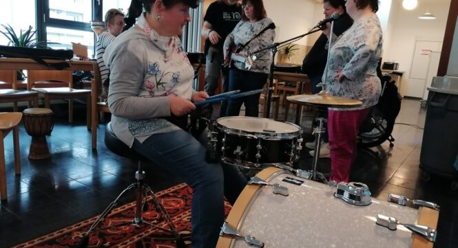 eine Frau sitzt am Schlagzeug , im Hintergrund mehrere Personen mit Instrumenten