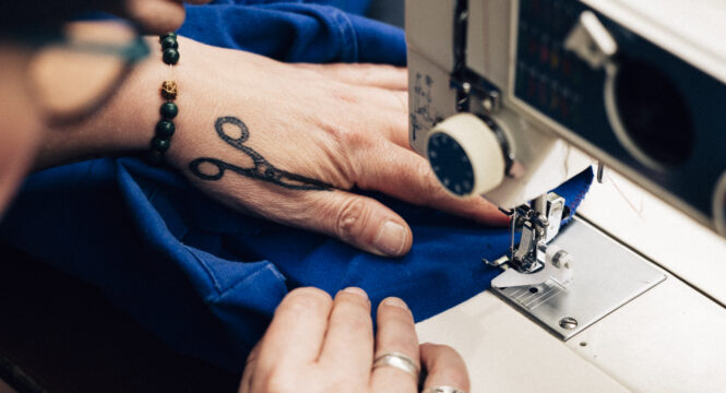 Buntfoto von einer Nähmaschine die von den Händen einer Frau bedient wird, die ein Tattoo von einer Schere auf der Hand hat