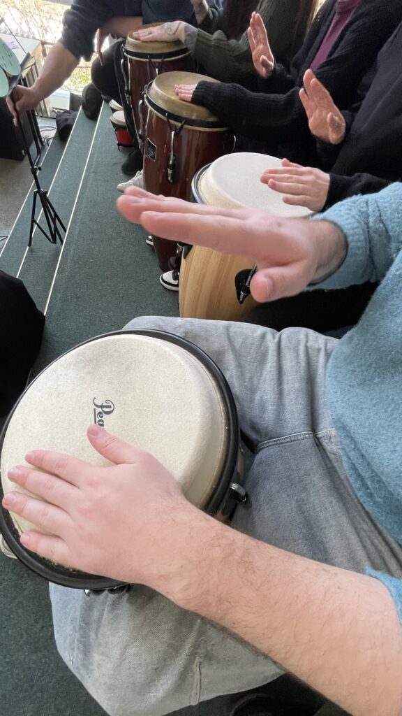 mehrere Menschen sitzen nebeneinander, man sieht Hände, die auf Congas (große Fasstrommeln) schlagen