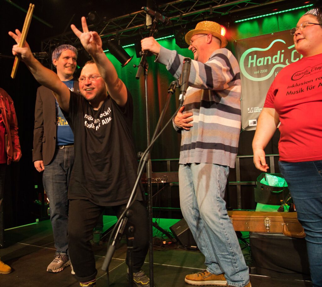 Vier erwachsene Personen unterschiedlichen Alters stehen auf einer Bühne und machen Musik. Eine Person reißt die Arme hoch und macht mit einer Hand ein V. In der anderen Hand sind Schlagzeug-Sticks. Auf zwei T-Shirts steht "Inklusion rockt!" Alle vier Personen sehen fröhlich aus.