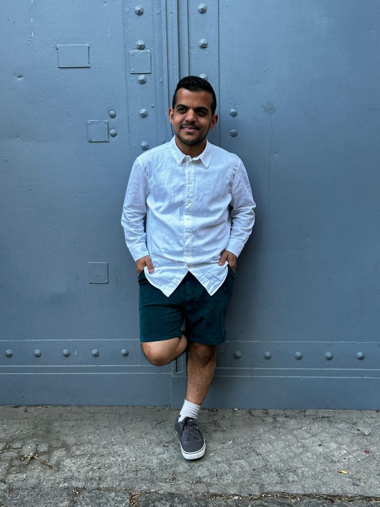 Eine Person steht locker an eine graue Metalltür gelehnt. Sie trägt ein weißes Hemd und eine kurze dunkelblaue Hose, dazu passend dunkelblaue Turnschuhe mit weißen Socken. Die Hände sind locker in die Hosentaschen gesteckt.