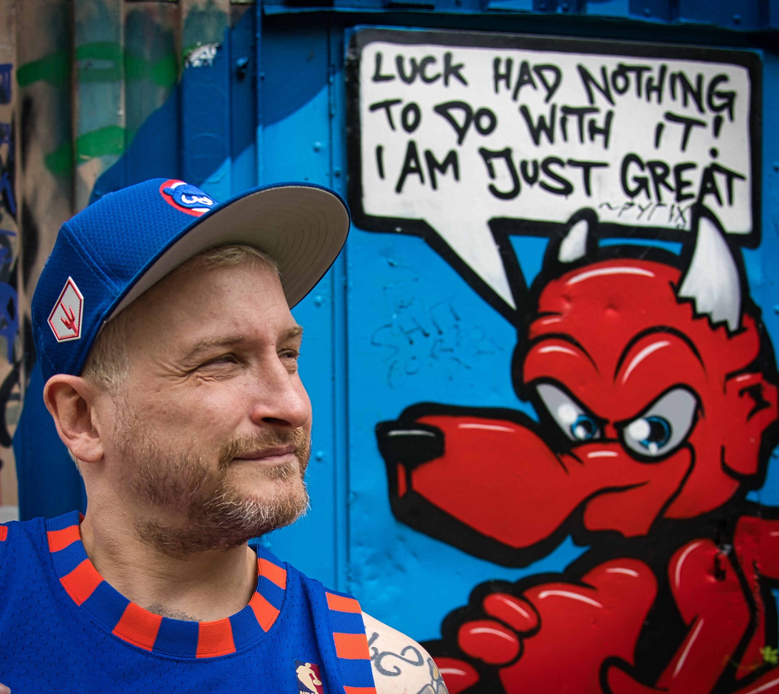 Buntfoto von einem Mann mit blauer Basecap, im HIntergrund ein Grafity, mit einem Teufel mit Sprechblase "Luck had nothing to do with it. I'm just great."