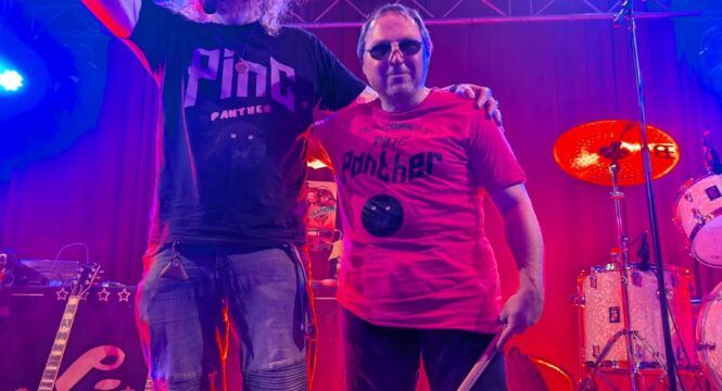 Zwei Personen stehen auf einer Bühne in rot-lila Licht. Sie schauen lachend und winkend in die Kamera. Beide tragen T-Shirts und Jeans. Neben ihnen sind Instrumente wie Gitarren und Schlagzeug zu sehen.