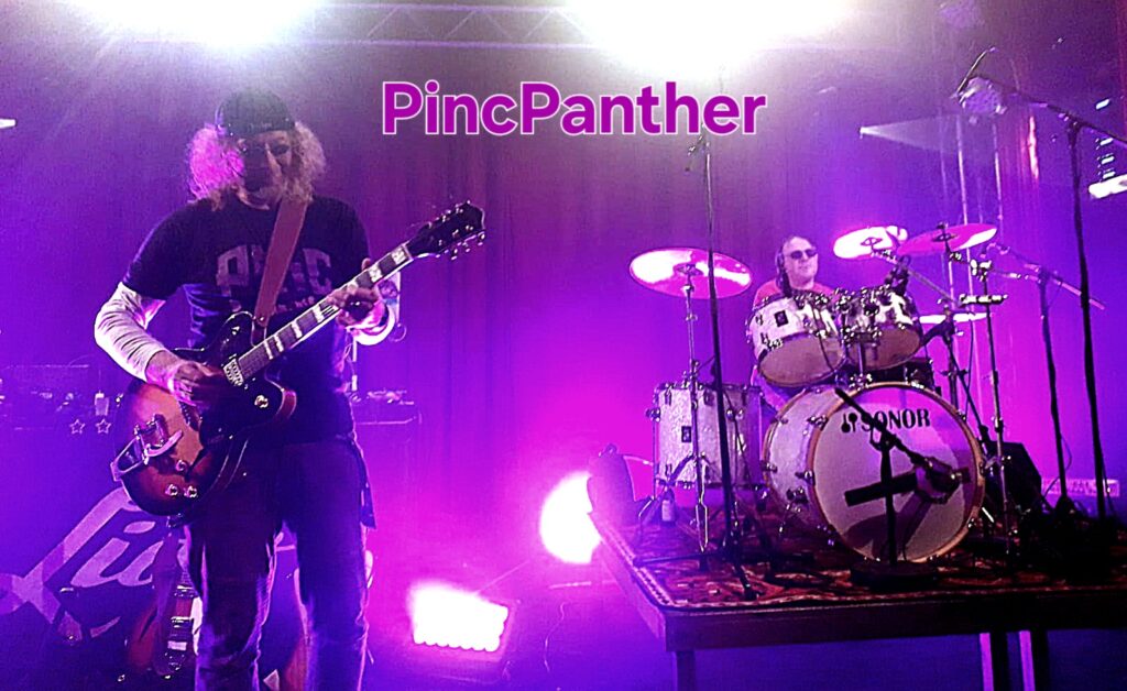 Eine Bühne im lila Licht. Im Vordergrund eine Person mit Locken und Mütze an einer E-Gitarre. Im Hintergrund rechts im Bild ist eine Schlagzeugspielende Person zu sehen. Beide Personen haben Sonnenbrillen auf.