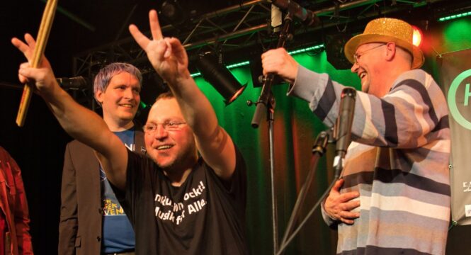 Drei Männer fröhlich nach einem Auftritt auf der Bühne, einer hält Schlagzeugstöcke hoch und macht ein Victory Zeichen ins Publikum.